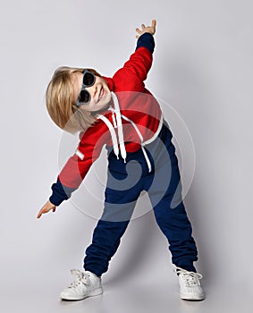 Smiling active frolic blond kid boy in blue and red hoodie, pants and sunglasses does gym exercises