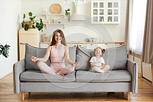 Smiling active young mom and little daughter sit on couch practicing yoga in lotus position