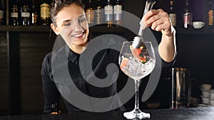 Smiling adorable female bartender putting strawberry in a glass with crushed ice while preparing cocktail. Nightlife in