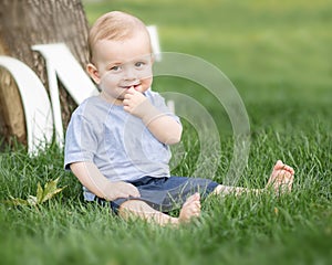 A smiling adorable small baby boy holding a finger in his mouth sitting on green grass outdoor at summer park. Emotions, smile, su