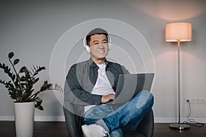 Smiling adult asian man in wireless headphones enjoy favorite music, typing on laptop, watch video