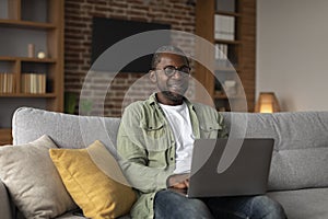 Smiling adult black male in glasses watch funny video on pc, chatting, surfing in internet, sits on sofa