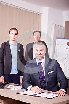 Smiling adult businessman at the desk and his younger colleagues