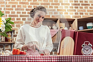 smiling adult housewife in vintage clothes cutting tomato and looking at camera