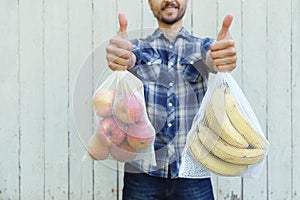 Smiling adult man holding reusable eco bags with fresh fruits and showing LIKE sign. Zero waste shopping, ban single use plastic