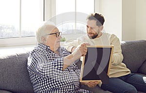 Smiling adult son sitting on sofa at home with senior father and looking to family photograph.