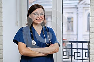 Smiling adult woman doctor in blue uniform stethoscope with folded arms, confident female medic looking at camera