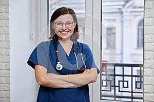 Smiling adult woman doctor in blue uniform stethoscope with folded arms, confident female medic looking at camera, standing near