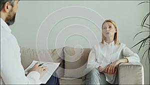 Smiling adult woman sitting on couch talking to male psychologist or psychoanalyst in office