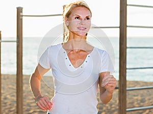 Smiling adult woman in white T-shirt is jogging on the beach