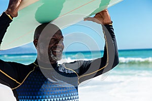 Smiling african american bald retired senior man carrying surfboard on head at beach