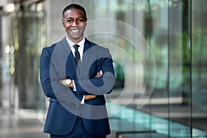 Smiling african american businessman CEO standing proud with arms crossed outside office workplace, colorful, reflective glass bui