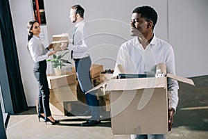 smiling african american businessman holding cardboard box while relocating with colleagues