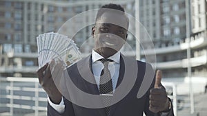 Smiling African American businessman showing money and thumb up at camera. Portrait of confident successful entrepreneur