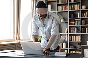 Smiling African American businessman standing near desk, using laptop