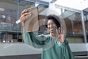 Smiling african american businesswoman waving on video call through tablet pc at workplace