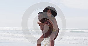 Smiling african american couple playing at the beach, man carrying woman piggyback