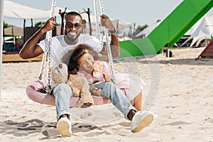 smiling african american father and daughter on spider web nest swing at amusement