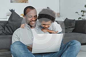 smiling african american father and son sitting on floor and using laptop