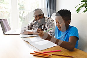 Smiling african american father and son sitting at table in dining room, working and doing homework