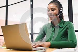 Smiling african-american female employee colleague using headset and laptop sitting on the workplace, talking on the