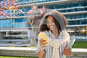Smiling African American girl student walking in park using mobile phone.