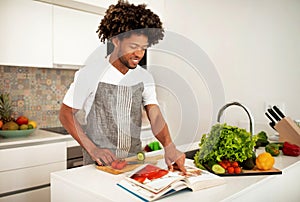 Smiling African American Guy Reading Paper Culinary Book In Kitchen