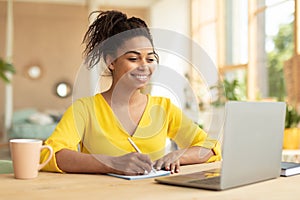 Smiling african american lady working on laptop taking notes in notebook, sitting at desk at home, copy space