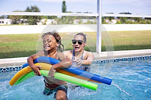 Smiling African American little boy playing in the swimming pool with his mother. Having fun playing with pool noodles in the sun