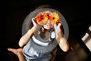 Smiling African American little girl closes eyes with colorful gerbera flowers in rays of sun on black background