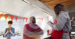 Smiling african american male food truck owner taking order from man, with woman using phone waiting