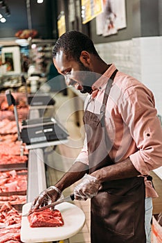 smiling african american male shop assistant in apron cutting steak of raw meat