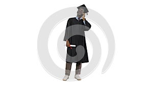 Smiling african american male student in graduation robe talking on the phone sharing happy news on white background.