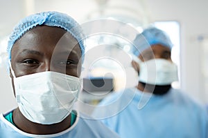 Smiling african american male surgeon in surgical mask and cap in operating theatre, copy space