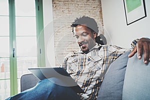 Smiling African American man in headphone making video call via electronic touch pad while sitting on sofa at home