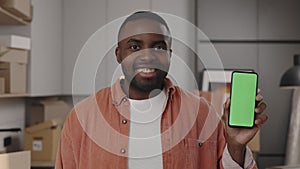 Smiling african american man holding smartphone and showing green screen phone to camera standing in new home. Mock up