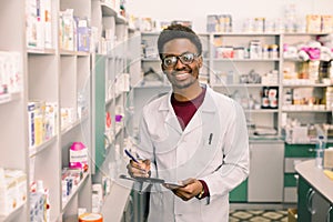 Smiling African American man pharmacist or Chemist Writing On Clipboard While standing in interior of pharmacy