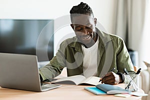 Smiling african american man sitting at desk, working on laptop and taking notes in notebook, black male studying online