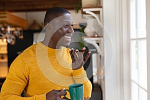 Smiling african american man in talking on smartphone holding coffee looking out of kitchen window