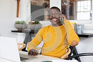 Smiling african american man using laptop at home and talking on smartphone