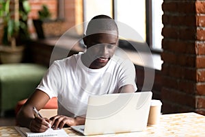 Smiling black male studying on laptop making notes in notebook