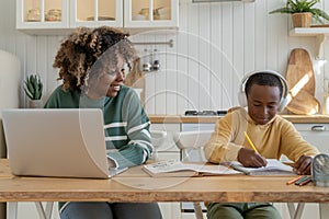 Smiling African American mother helping small son with homework while working remotely on laptop