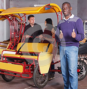 Smiling African-American pedicab driver standing near rickshaw cycle