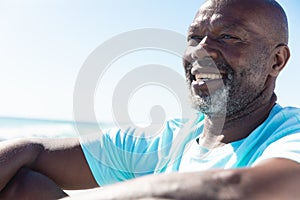 Smiling african american retired senior man with white beard sitting at beach on sunny day