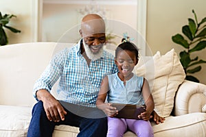 Smiling african american senior man with granddaughter using digital tablet while sitting on sofa