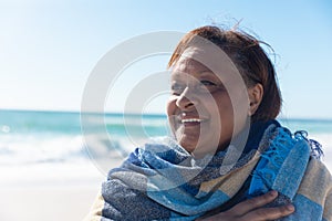 Smiling african american senior retired woman wrapped in shawl looking away at beach on sunny day