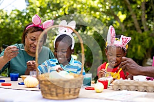 Smiling african american siblings and grandmother wearing bunny ears while painting eggs on easter