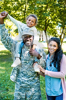 smiling african american soldier in military uniform holding daughter on shoulders