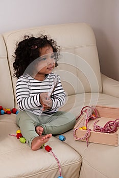 Smiling African American toddler with tousled curls plays with beads on a neutral couch, embodying innocence and