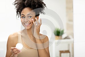Smiling african american woman applying facial moisturizer while holding jar and looking at mirror.